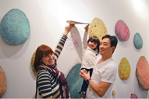 Two adults hold a toddler in their arms as they stand in front of an art installation in the ArtStarts Gallery