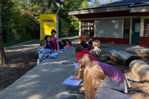 A group of children takes notes while an educator gives indication with the yellow side of Where Am I exhibit in the background