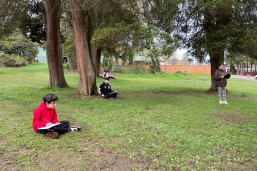 A group of children sitting and standing, spread out in a park taking notes.