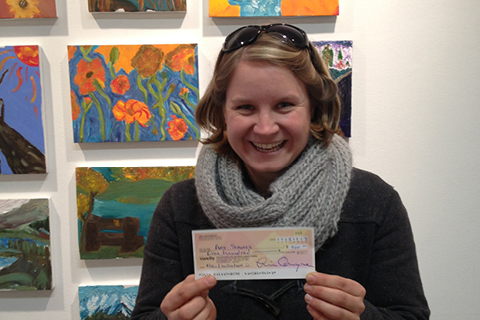 An adult stands in front of a series of artwork with a cheque in their hands