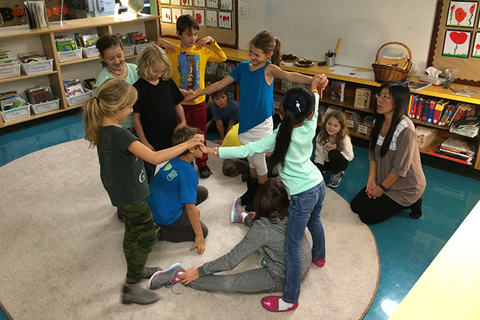 A group of young people and  an educator in a classroom setting. Several of the children are have their hands interlocked with o