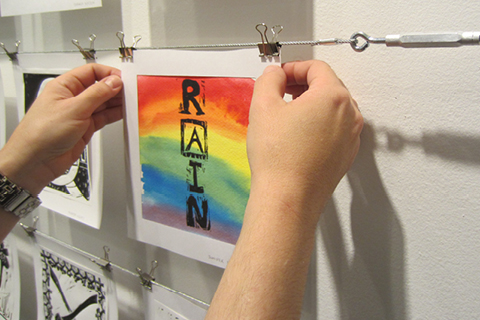 A person's hands holding a painting in rainbow colours with the letters 'RAIN' stamped on vertically