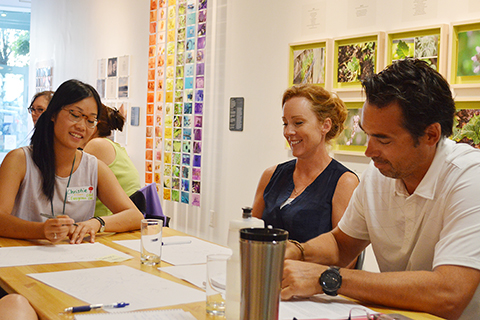 A group of three adults sit around a table and write on pieces of blank paper with mark making tools in the ArtStarts Gallery