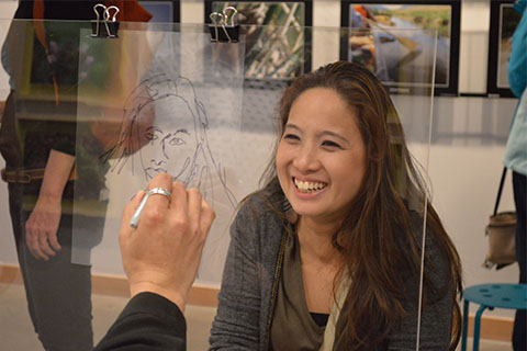 An adult smiles behind a clear plastic divider while someone traces their portrait