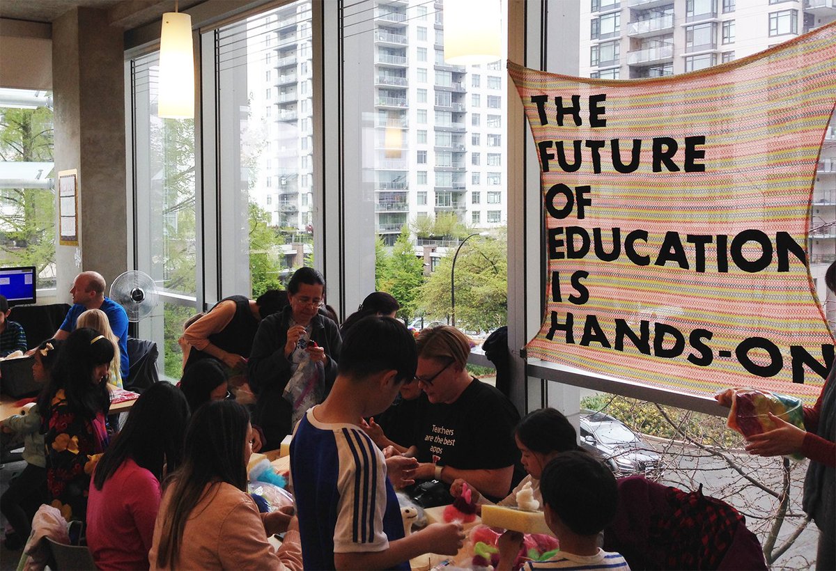 Younger learners and adults gathered around a table beside a woven banner that reads 'the future of education is hands on'